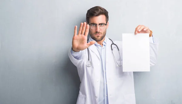 Handsome young doctor man over grey grunge wall holding blank sheet contract with open hand doing stop sign with serious and confident expression, defense gesture