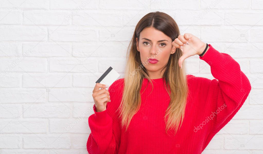 Young adult woman over white brick wall holding credit card with angry face, negative sign showing dislike with thumbs down, rejection concept