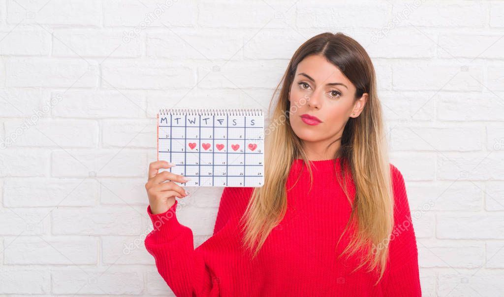 Young adult woman over white brick wall holding period calendar with a confident expression on smart face thinking serious
