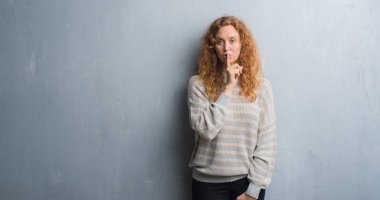 Dudaklarında parmak ile sessiz olmanı gri grunge duvarın üzerinden genç Kızıl saçlı kadın. Sessizlik ve gizli kavramı.