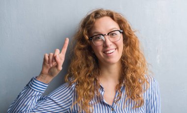 Genç Kızıl saçlı kadın gri grunge duvar bir fikir ya da soru işaret parmağı ile gülen yüz, sürpriz bir numara