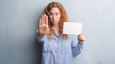 Gri grunge duvar holding boş kartı açık eliyle dur işareti ile ciddi ve kendine güvenen ifade, savunma hareketi yaparken genç Kızıl saçlı kadın