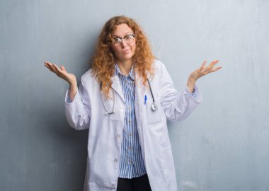 Genç redhead doktor kadın ceket clueless ve şaşkın ifade kollar ve eller kaldırdı ile giyen gri grunge duvarın üzerinden. Şüphe kavramı.