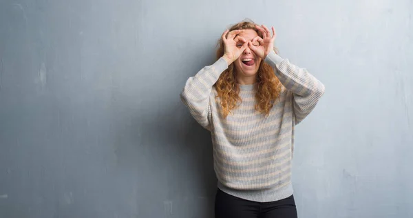 灰色垃圾墙上的年轻红头发的女人做 手势像望远镜伸出舌头 眼睛看通过手指 疯狂的表情 — 图库照片