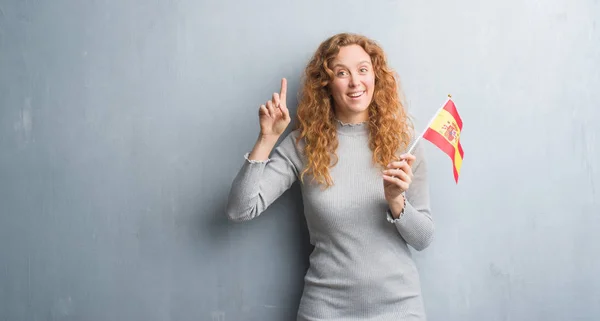 Joven Pelirroja Sobre Pared Grunge Gris Sosteniendo Bandera España Sorprendida —  Fotos de Stock