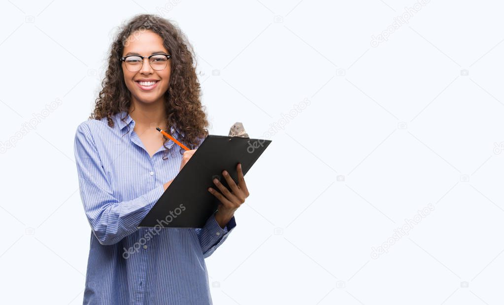 Young hispanic business woman holding clipboard with a happy face standing and smiling with a confident smile showing teeth