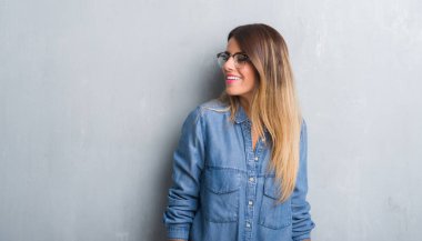 Grunge Genç Yetişkin kadın uzak tarafına yüzünde, doğal ifade gülümseme ile seyir gözlüklü duvar gri. Kendine güvenen gülüyor.