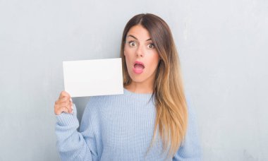 Gri grunge duvar holding boş kartı bir sürpriz yüz, korkuyor ve heyecanlı korku ifadesi ile şok korkmuş Genç Yetişkin kadın