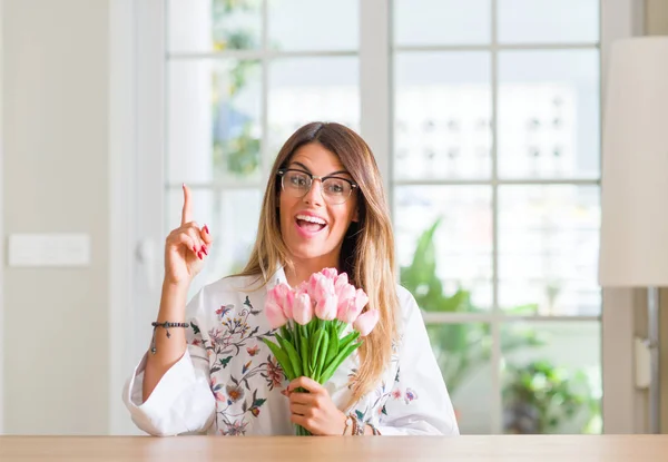Young woman at home holding pink tulips flowers surprised with an idea or question pointing finger with happy face, number one