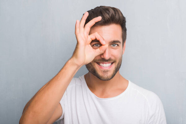 Handsome young man over grey grunge wall with happy face smiling doing ok sign with hand on eye looking through fingers