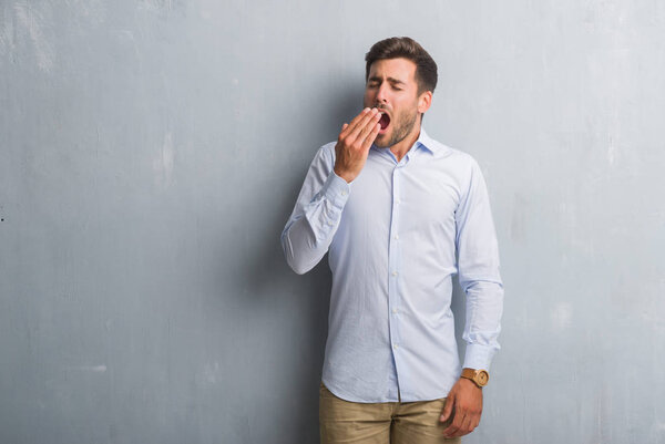 Handsome young business man over grey grunge wall wearing elegant shirt bored yawning tired covering mouth with hand. Restless and sleepiness.