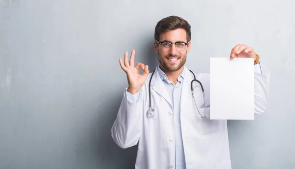 Handsome young doctor man over grey grunge wall holding blank sheet contract doing ok sign with fingers, excellent symbol