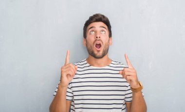 Yakışıklı delikanlı lacivert t-shirt giyen gri grunge duvarın üzerinden hayran ve yukarıya bakıyor ve parmakları ile işaret ve silah kaldırdı şaşırttı.