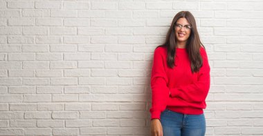 Genç esmer kadın beyaz tuğla duvarın üzerinde duruyor kameraya seksi bir ifadeyle bakıyor, neşeli ve mutlu bir yüzle..