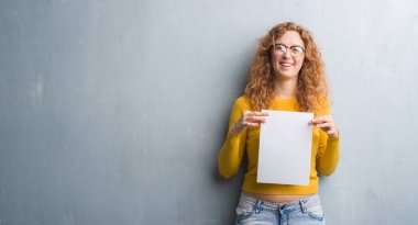 Gri grunge duvar boş kağıt sayfası ile ayakta ve diş gösterilen kendine güvenen bir gülümsemeyle gülümseyerek mutlu bir yüz tutan genç Kızıl saçlı kadın