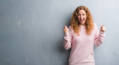 Genç redhead gri grunge duvar giyen üzerinde pembe kadın kutluyor kazak şaşırttı ve silah ile başarı için hayran kaldırdı ve gözleri açık. Kazanan kavramı.