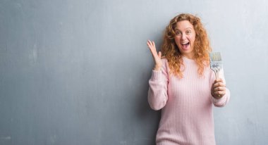 Gri grunge duvar boya tutan genç Kızıl saçlı kadın çok mutlu ve heyecanlı, kazanan ifade büyük gülümseme ile çığlık zaferini kutluyor fırça ve ellerini kaldırdı