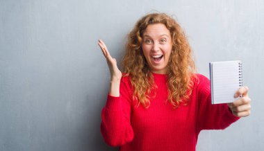 Gri grunge genç Kızıl saçlı kadın duvar büyük gülümseme ile çığlık zaferini kutluyor ve ellerini kaldırdı boş defter çok mutlu ve heyecanlı, kazanan ifade gösterilen