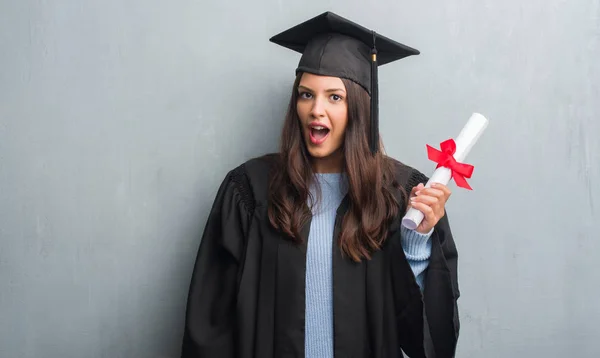若いブルネットの女性グランジに灰色の驚き顔 恐怖表現と興奮とショックで怖い学位を保持する大学院の制服を着て壁 — ストック写真