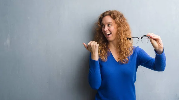 Young Redhead Woman Grey Grunge Wall Holding Optical Glasses Pointing — Stock Photo, Image