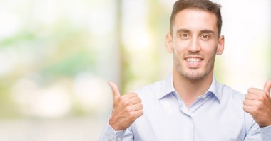 Yakışıklı genç işadamı başarı işareti başparmak gülümseyen ve mutlu el ile olumlu hareketi yapıyor. Neşeli bir ifadeyle, kazanan jest kameraya bakıyor.