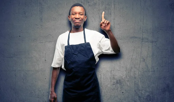 African man shop owner wearing apron raising finger, the number one