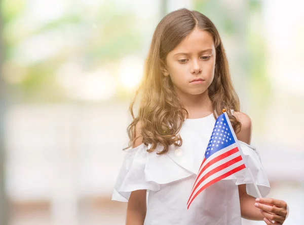 Brunette hispanic girl holding flag of United States of America with a confident expression on smart face thinking serious