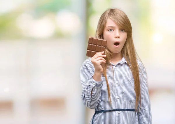 Young Blonde Child Holding Chocolate Bar Scared Shock Surprise Face — Stock Photo, Image