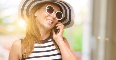 Orta yaş serin yaz şapka ve güneş gözlüğü smartphone cep telefonuyla konuşurken mutlu giyen kadın