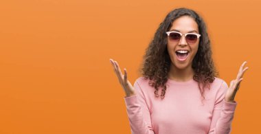 Beautiful young hispanic woman wearing sunglasses celebrating mad and crazy for success with arms raised and closed eyes screaming excited. Winner concept clipart
