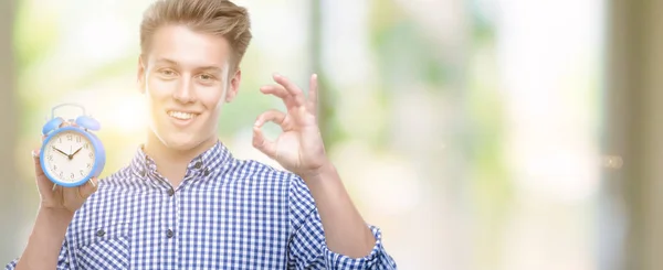 Young Handsome Blond Man Holding Alarm Clock Doing Sign Fingers — Stock Photo, Image
