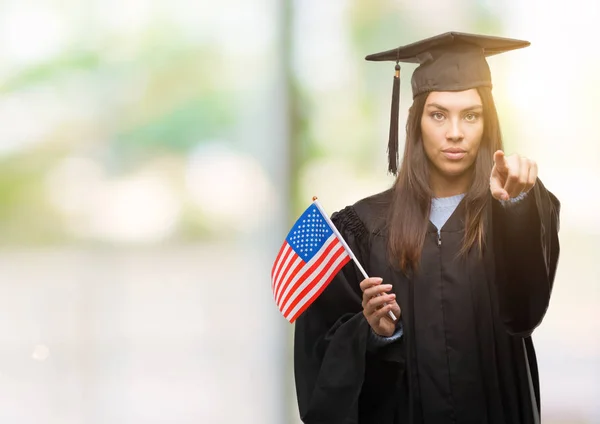 Young hispanic woman wearing graduated uniform holding flag of america pointing with finger to the camera and to you, hand sign, positive and confident gesture from the front