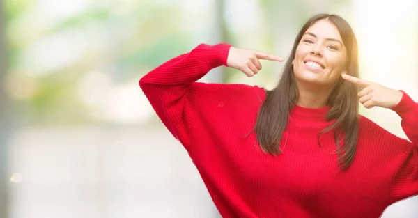 年轻漂亮的西班牙裔穿着红色毛衣微笑着自信地显示和指着手指的牙齿和嘴巴 健康理念 — 图库照片