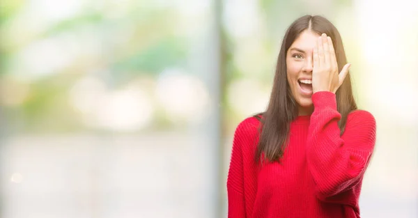 年轻漂亮的西班牙裔穿着红色毛衣覆盖一只眼睛与自信的脸上的微笑和惊喜的情绪 — 图库照片