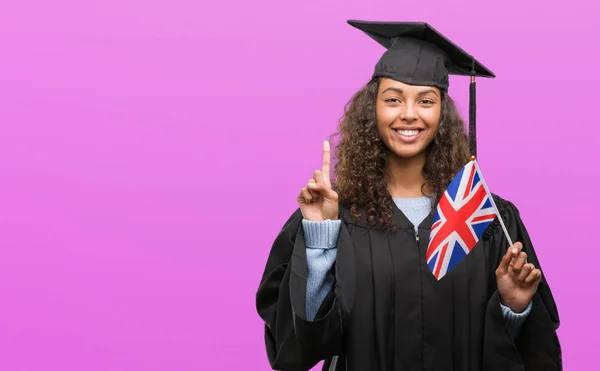 Young hispanic woman wearing graduation uniform holding flag of UK surprised with an idea or question pointing finger with happy face, number one