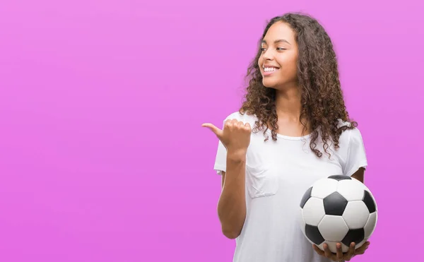 年轻的西班牙裔女子拿着足球球指着和显示与拇指与快乐的脸微笑的一面 — 图库照片