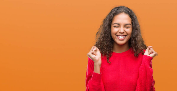 Young hispanic woman wearing red sweater excited for success with arms raised celebrating victory smiling. Winner concept.
