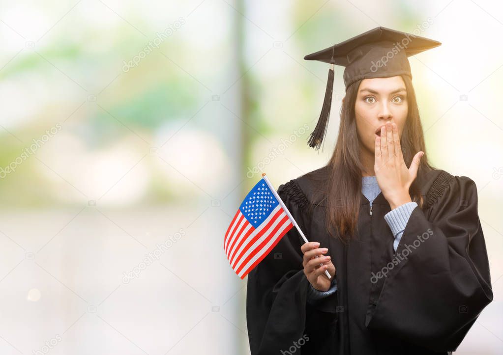 Young hispanic woman wearing graduated uniform holding flag of america cover mouth with hand shocked with shame for mistake, expression of fear, scared in silence, secret concept