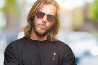 Genç yakışıklı adam uzun saçlı izole arka plan şüpheci ve sinir, onaylamayan ifade yüz çapraz kolları üzerinde güneş gözlüğü takıyor. Negatif kişi.