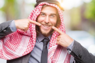 Genç yakışıklı Arap adam uzun saçlı emin gösterilen ve işaret parmakları diş ve ağız ile gülümseyen izole arka plan üzerinde keffiyeh giyiyor. Sağlık kavramı.