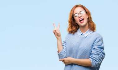 İzole arka plan mutlu yüz zafer işareti yaparken kameraya göz kırpıyor ile gülümseyen kış kazak giyen genç güzel kadın. İki numaralı.