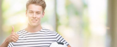 Genç yakışıklı sarışın adam futbol topu ok işareti, başparmak ile parmak, mükemmel işareti yapıyor kocaman bir gülümseme ile mutlu holding