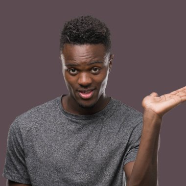 Neşeli sunulması ve kameraya bakarak avucunun ile işaret gülümseyen gri t-shirt giyen genç Afro-Amerikan adam.