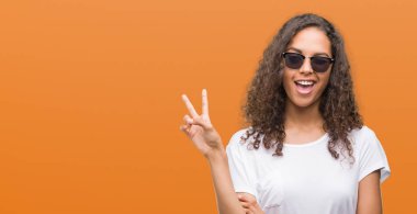 Genç İspanyol kadın mutlu yüz zafer işareti yaparken kameraya göz kırpıyor ile gülümseyen güneş gözlüğü takıyor. İki numaralı.