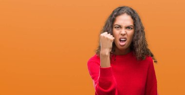 Genç İspanyol kadın giyiyor kırmızı kazaklı öfkeli ve sinirli sinirli ve öfke ile bağırarak süre öfkeli yumruk yükselterek. Öfke ve agresif kavramı.