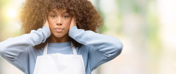 African american shop owner woman wearing an apron covering ears ignoring annoying loud noise, plugs ears to avoid hearing sound. Noisy music is a problem.