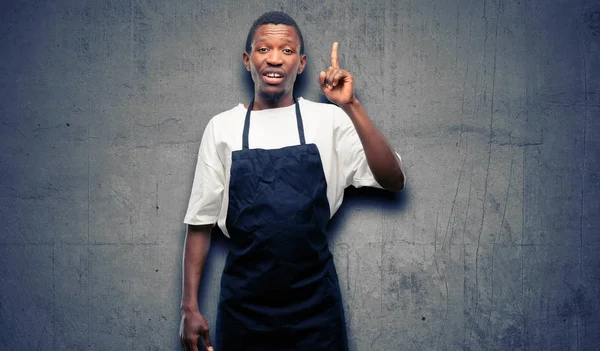 African man shop owner wearing apron happy and surprised cheering expressing wow gesture pointing up