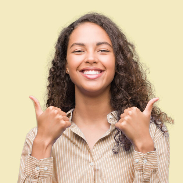 Young hispanic business woman success sign doing positive gesture with hand, thumbs up smiling and happy. Looking at the camera with cheerful expression, winner gesture.