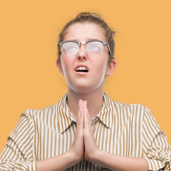 Young blonde business woman begging and praying with hands together with hope expression on face very emotional and worried. Asking for forgiveness. Religion concept.