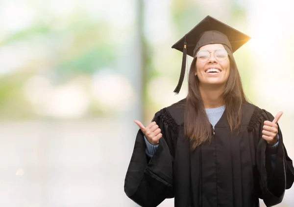年轻的西班牙裔妇女穿着毕业帽和一致的成功标志做积极的手势与手 大拇指向上微笑和快乐 看着带着欢快表情的相机 胜利者的手势 — 图库照片